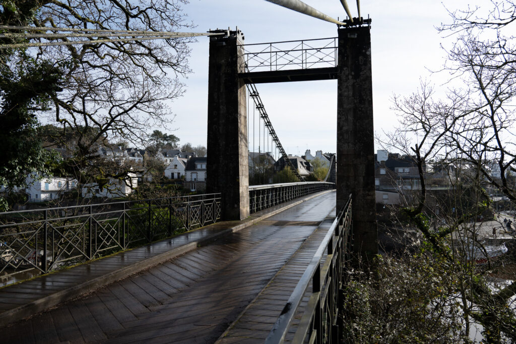 Un pont piétonnier en bois, doté de garde-corps métalliques et d'un haut pilier en pierre, enjambe une rivière, avec des maisons et des arbres visibles en arrière-plan par une journée nuageuse.