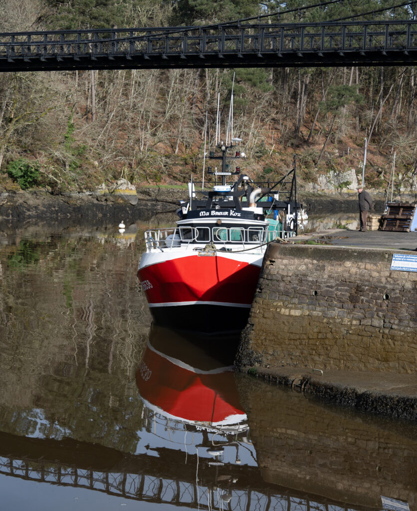 Un bateau rouge et blanc amarré à un quai en pierre au bord d'une rivière, dont il se reflète dans l'eau. Une passerelle noire enjambe le cours d'eau, tandis qu'à l'arrière-plan, des arbres bordent la rive.
