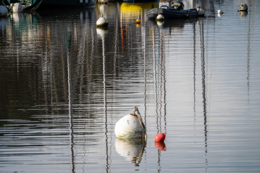 Des bouées flottent sur une eau calme, reflétant les bateaux et les structures environnants. La surface de l'eau est ondulée, ce qui déforme les reflets. Une grande bouée blanche et une bouée rouge plus petite se détachent au premier plan.