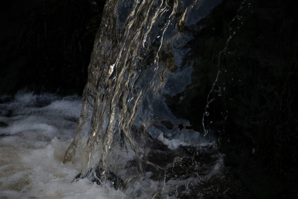 Une cascade d'eau limpide dévale un rocher sombre, formant de l'écume blanche à sa base. L'eau semble scintiller, la lumière se reflétant sur sa surface et mettant en valeur son mouvement et sa texture. L'arrière-plan est plongé dans l'ombre et contraste avec la clarté de l'eau.