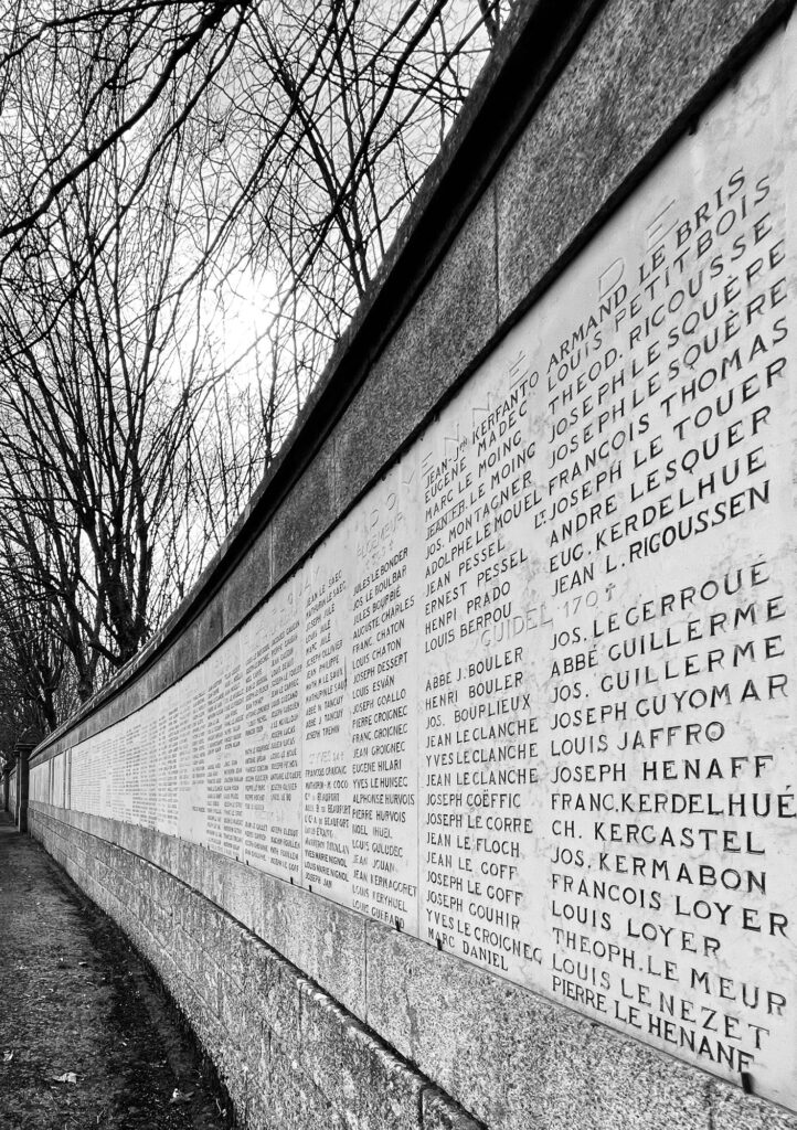 Une photo en noir et blanc montrant un mur commémoratif en pierre incurvé, sur lequel sont gravés des noms, situé le long d'une allée bordée d'arbres dénudés, sous un ciel couvert.