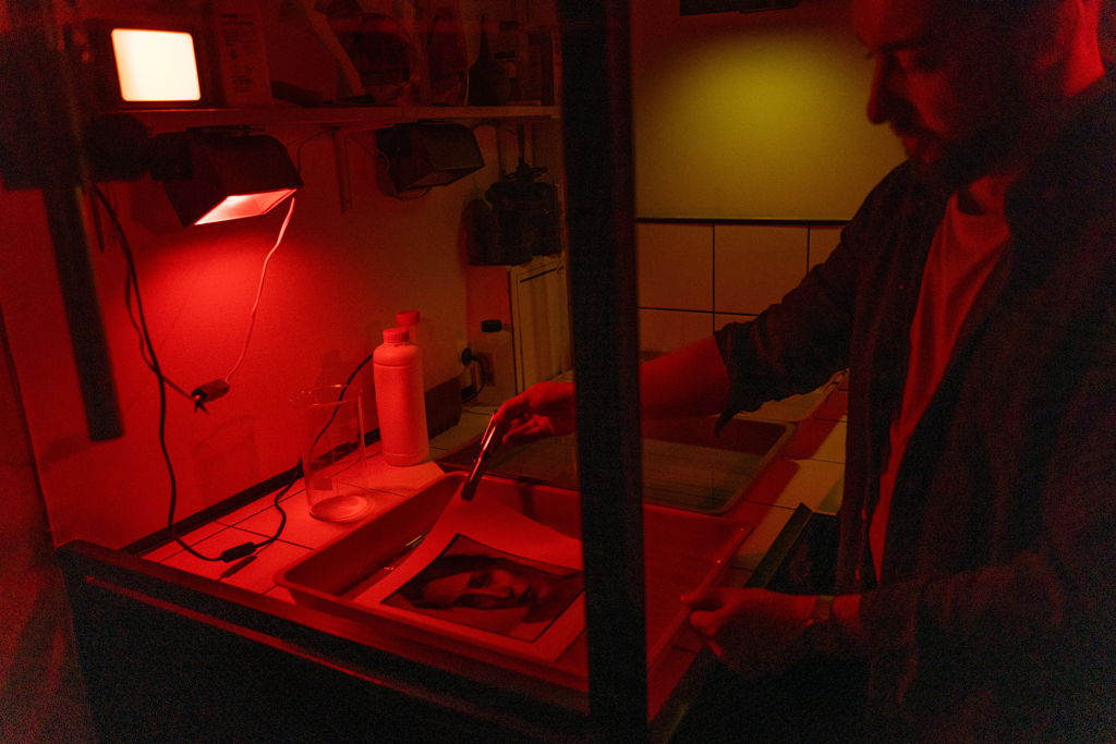 Une personne développe une photo en noir et blanc sous une lumière rouge dans une chambre noire, en utilisant des pinces pour déplacer le tirage dans un bac de traitement. Du matériel photographique et des flacons sont visibles sur les étagères à proximité.